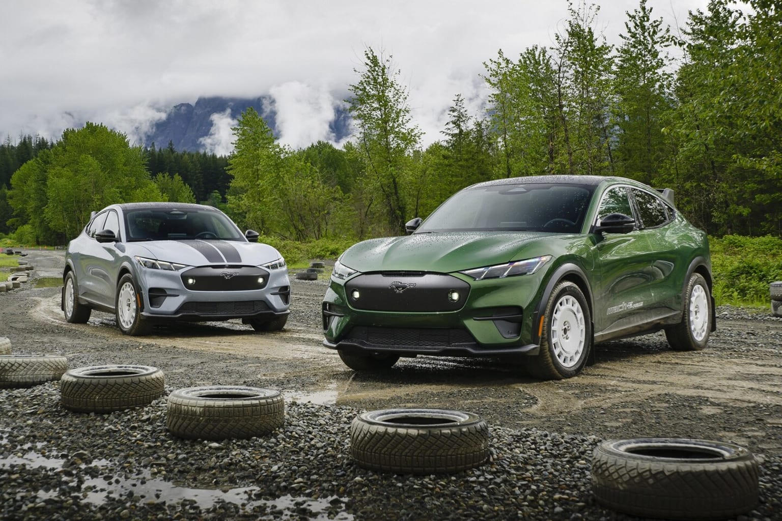 Two sleek, modern SUVs parked on a gravel road surrounded by lush, green trees and a cloudy, mountainous backdrop.