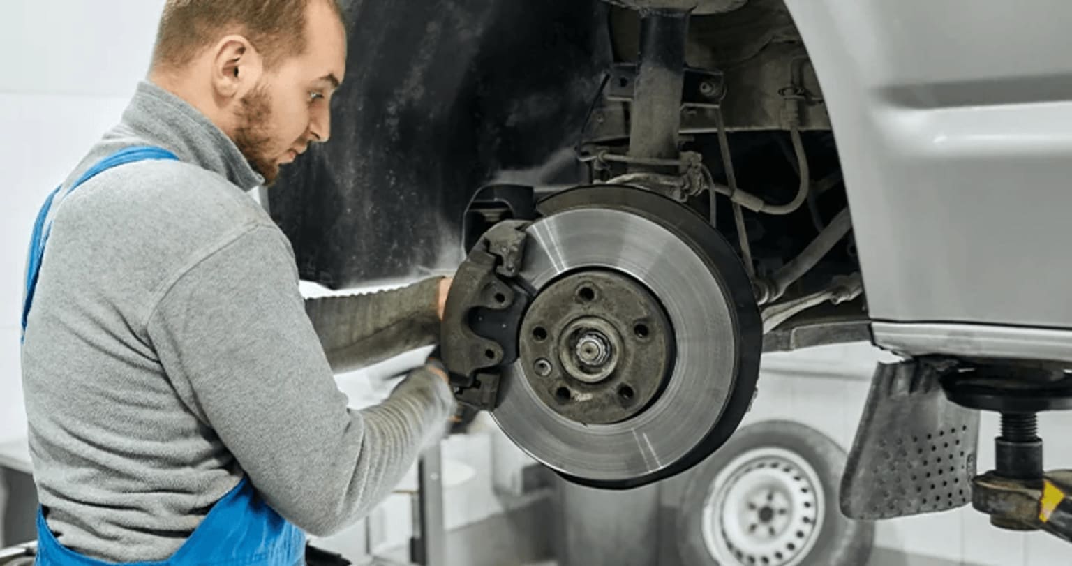 A mechanic in a gray sweater is performing brake maintenance on a vehicle, inspecting the brake disc and various components in the foreground.