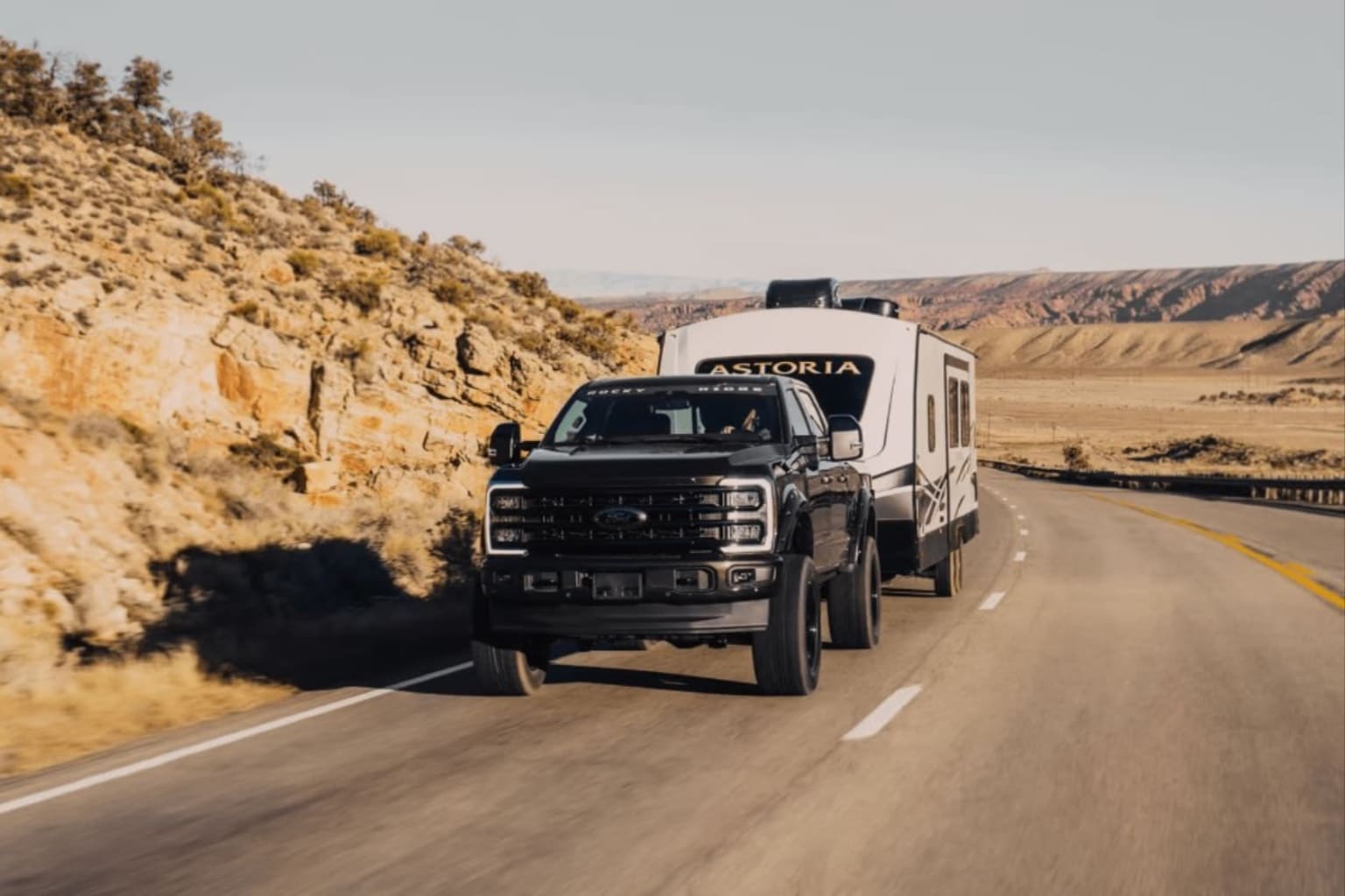A black truck tows a white travel trailer down a paved road through a desert landscape with rocky hills.