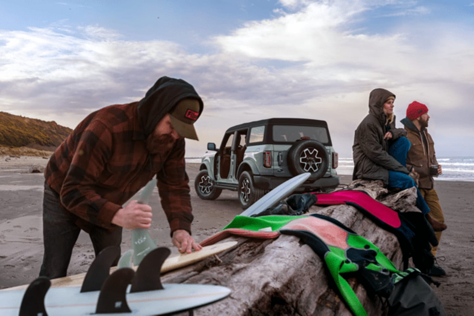 People in warm clothing standing on a beach with a vehicle in the background and a surfboard in the foreground.