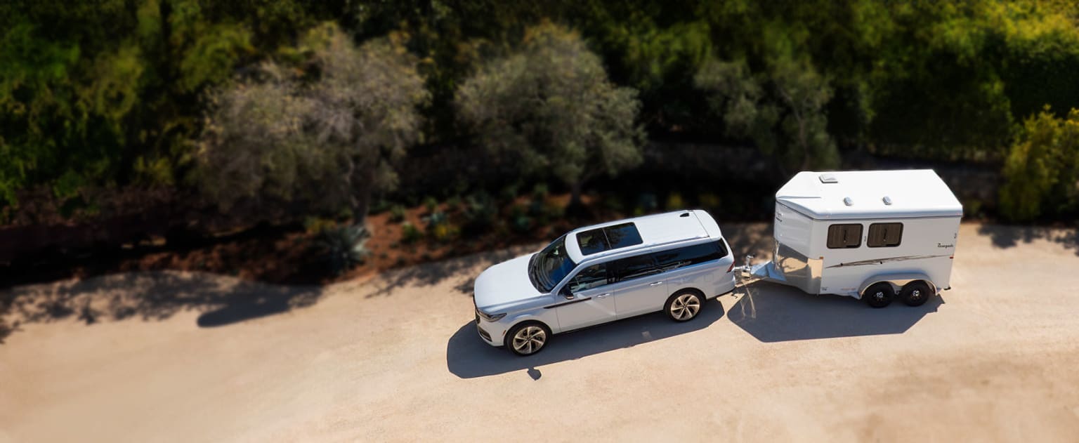 A white SUV towing a small camper trailer parked on a dirt road surrounded by lush, green foliage.