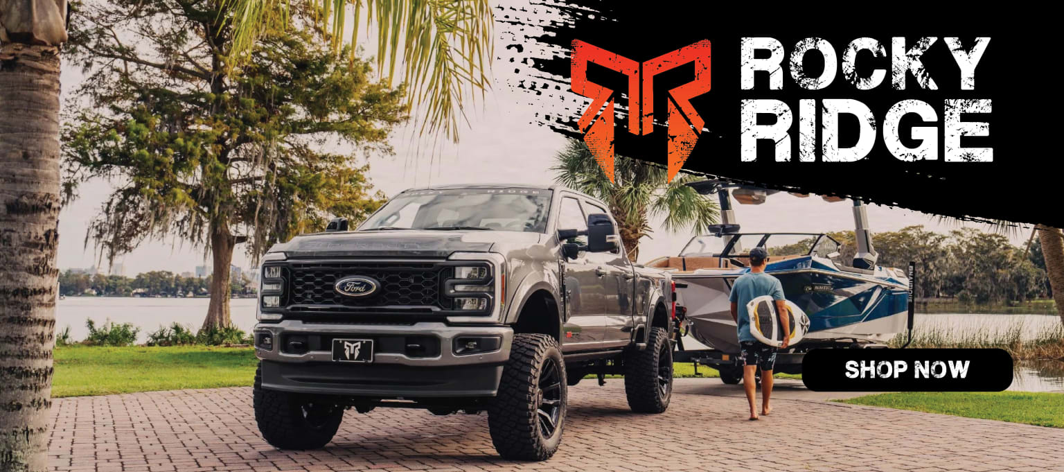 A large, lifted truck with a logo prominently displayed stands in the foreground, surrounded by palm trees and a body of water in the background.