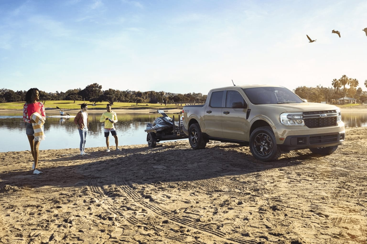 A group of people standing near a large silver pickup truck on a sandy beach, with a body of water and trees in the background.