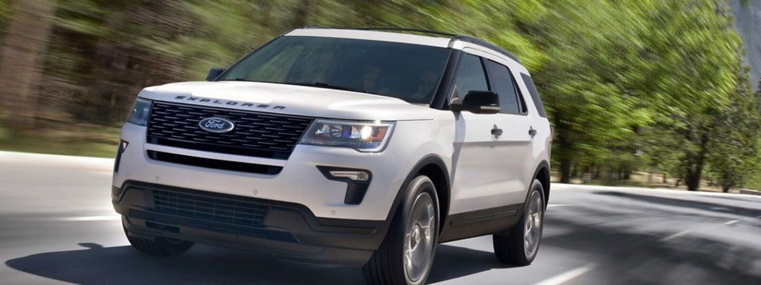 a white ford explorer driving down a road next to a forest of green trees on a sunny day with a blurry image of the front end of the car