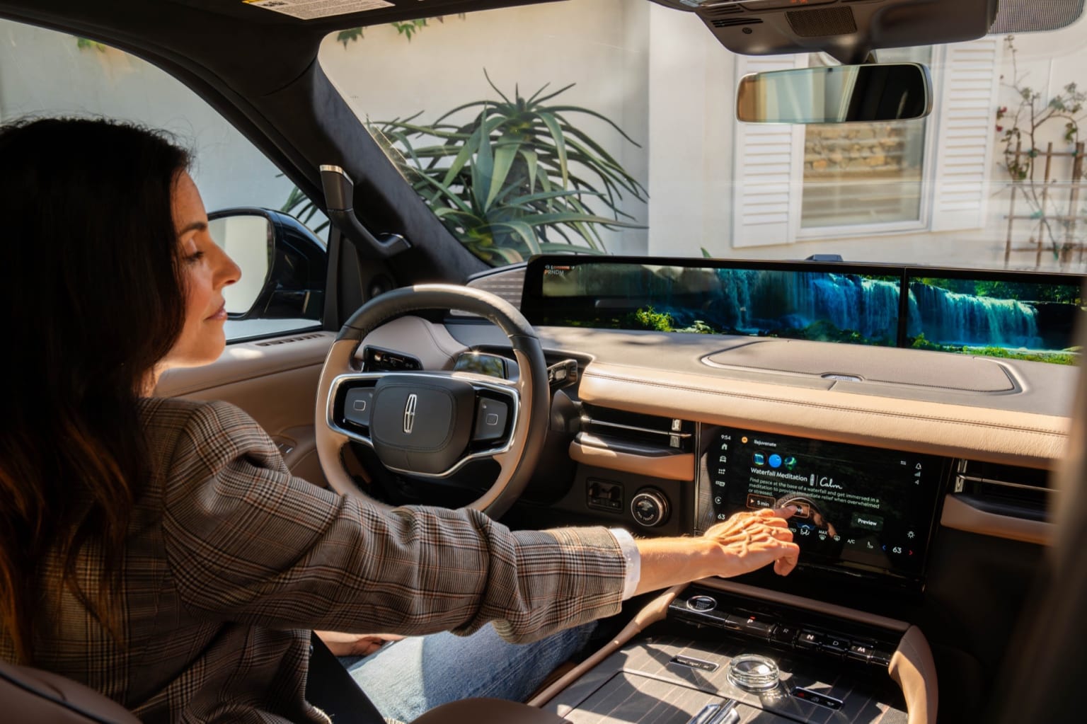 A person sitting in the driver's seat of a vehicle, interacting with the controls and display while a plant is visible in the background.