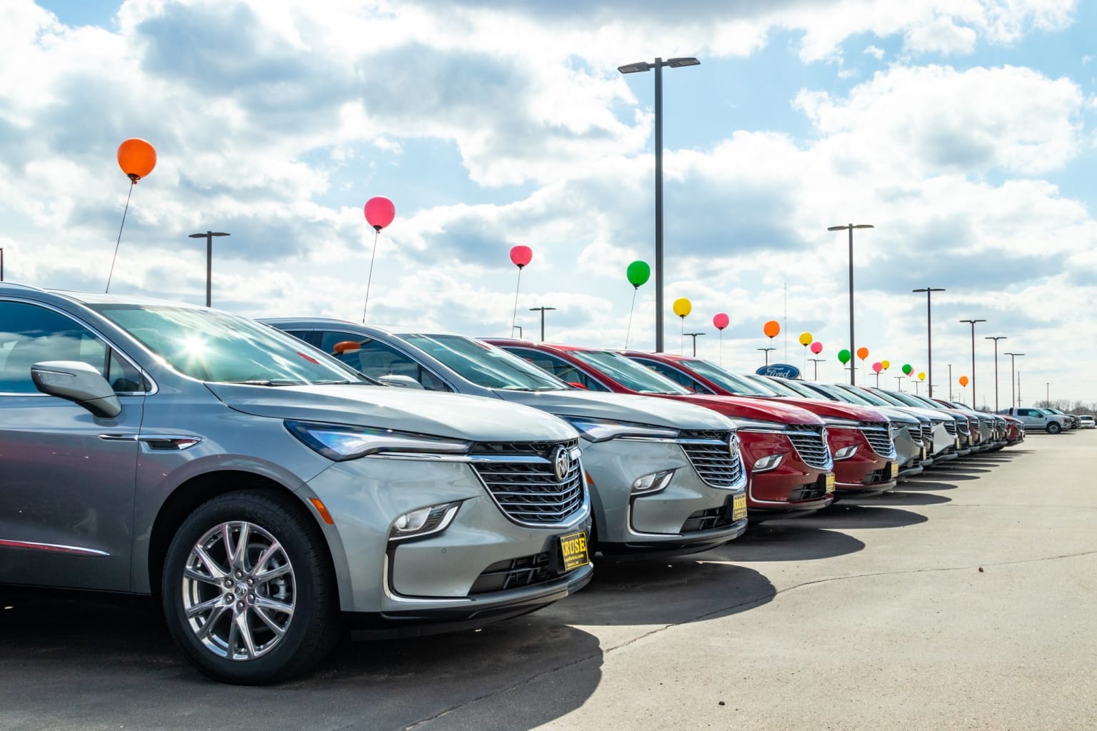 a row of cars parked in a parking lot with balloons in the sky in the back of the row