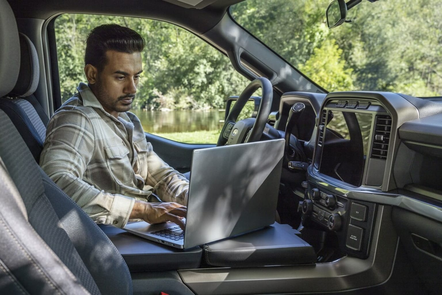 A person in the driver's seat of a vehicle, using a laptop computer with a natural, wooded environment outside.
