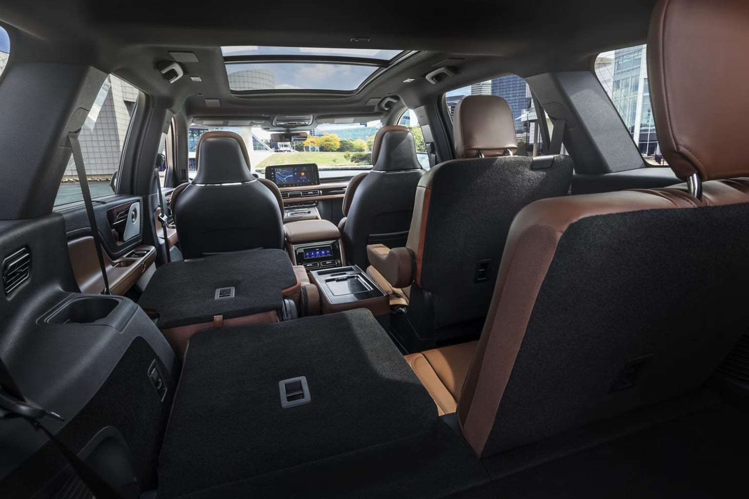 Interior of a spacious vehicle with multiple rows of seats, panoramic sunroof providing natural light. Seats upholstered in a combination of dark and light colors creating a visually appealing contrast. Urban setting background with buildings and other structures visible through the windows.