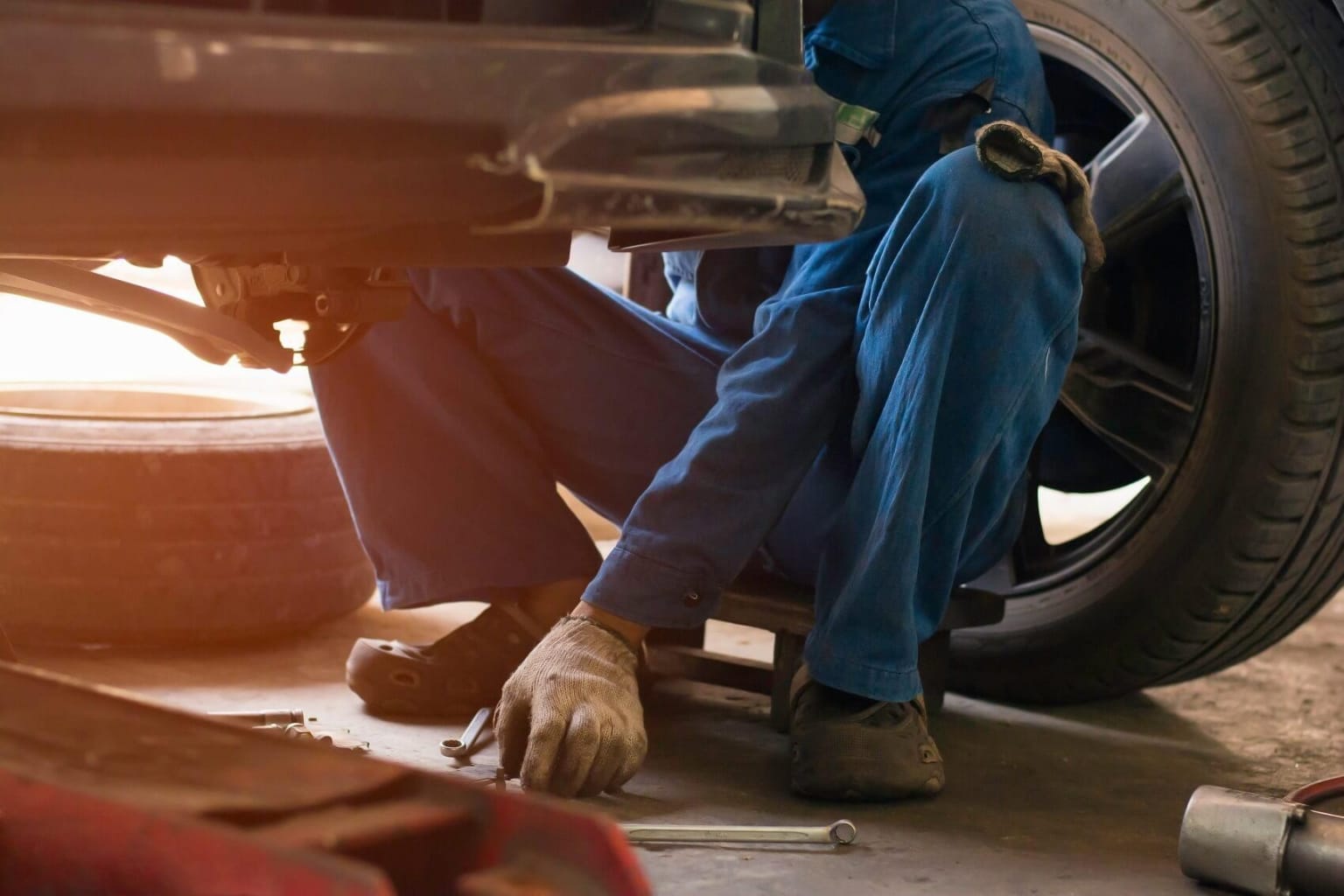 An automotive technician in a blue coverall is servicing a vehicle, surrounded by various tools and equipment in the background.