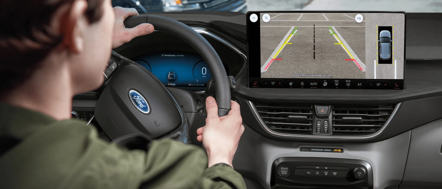 Interior of a vehicle with driver's hands on the steering wheel and a display screen showing a rearview camera view.
