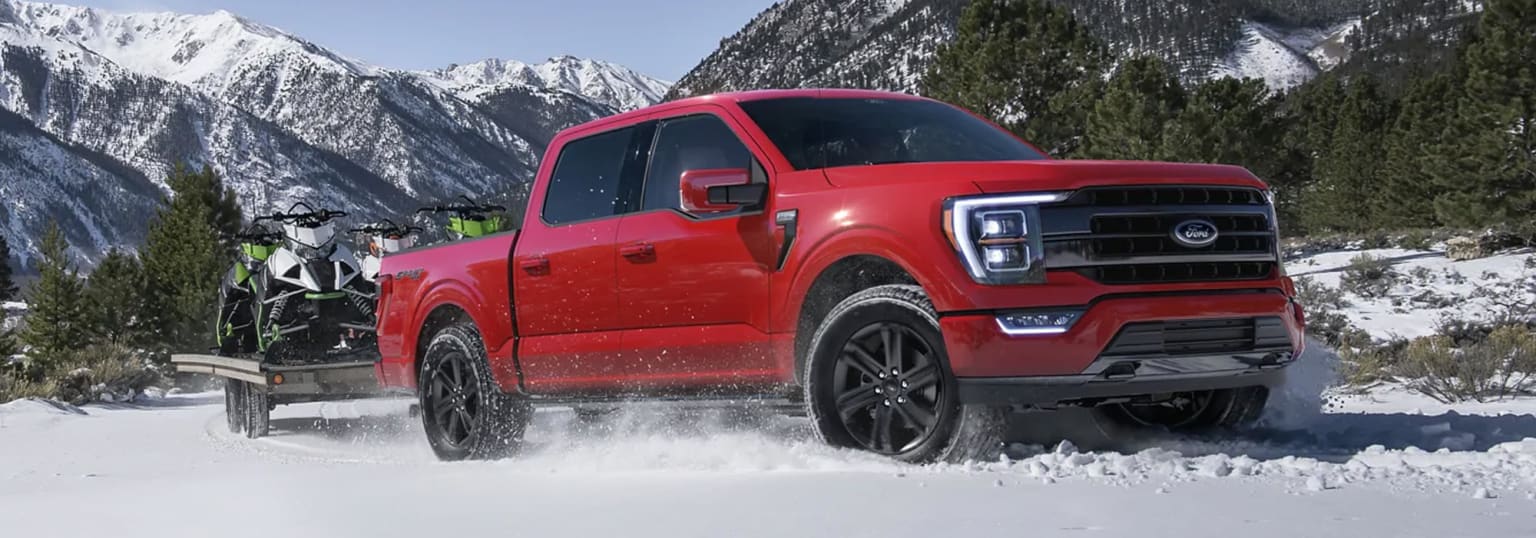 A red pickup truck driving through snow-covered terrain in front of a mountainous, snowy landscape.