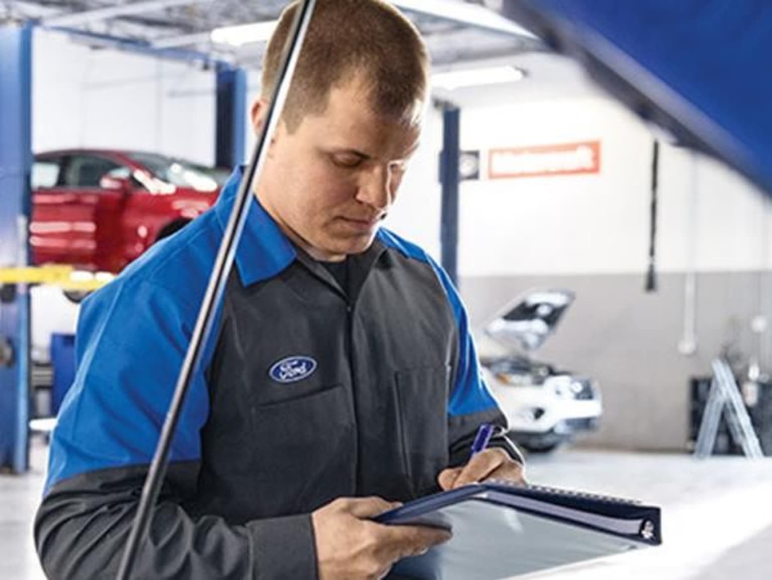 A mechanic in a uniform is using a tablet device while working on a car in a repair shop.