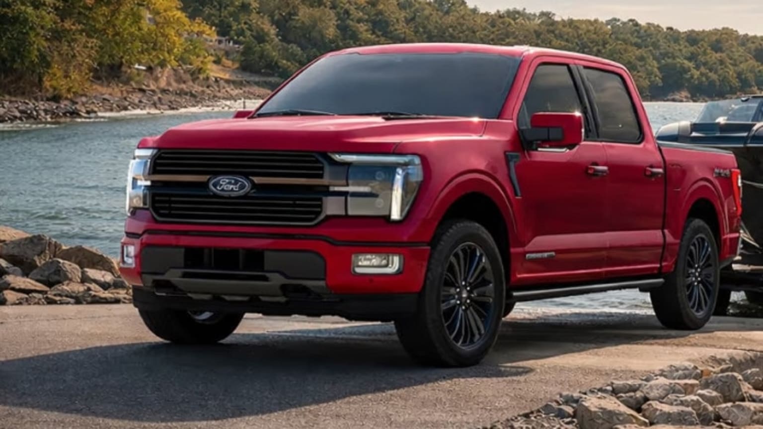 A red pickup truck is parked on a paved surface with a body of water and trees in the background.