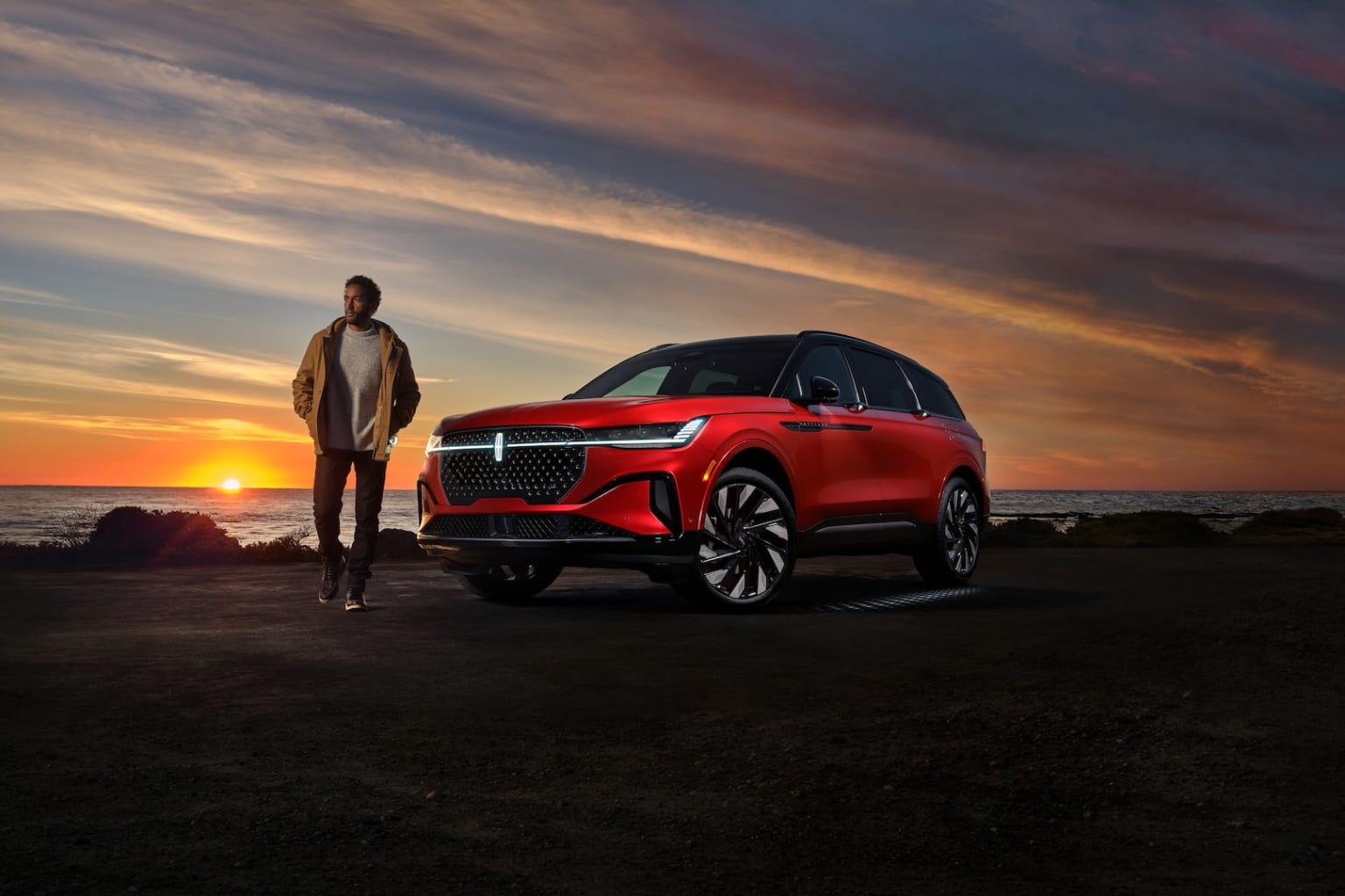 A person standing next to a red SUV-style vehicle against a dramatic sunset sky with colorful clouds.