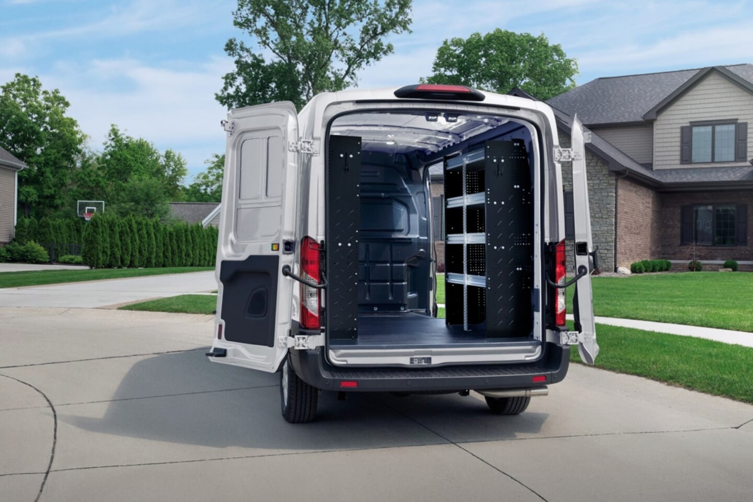 A white cargo van with its rear doors open, revealing shelving inside, sits in a driveway with a suburban house and trees in the background.