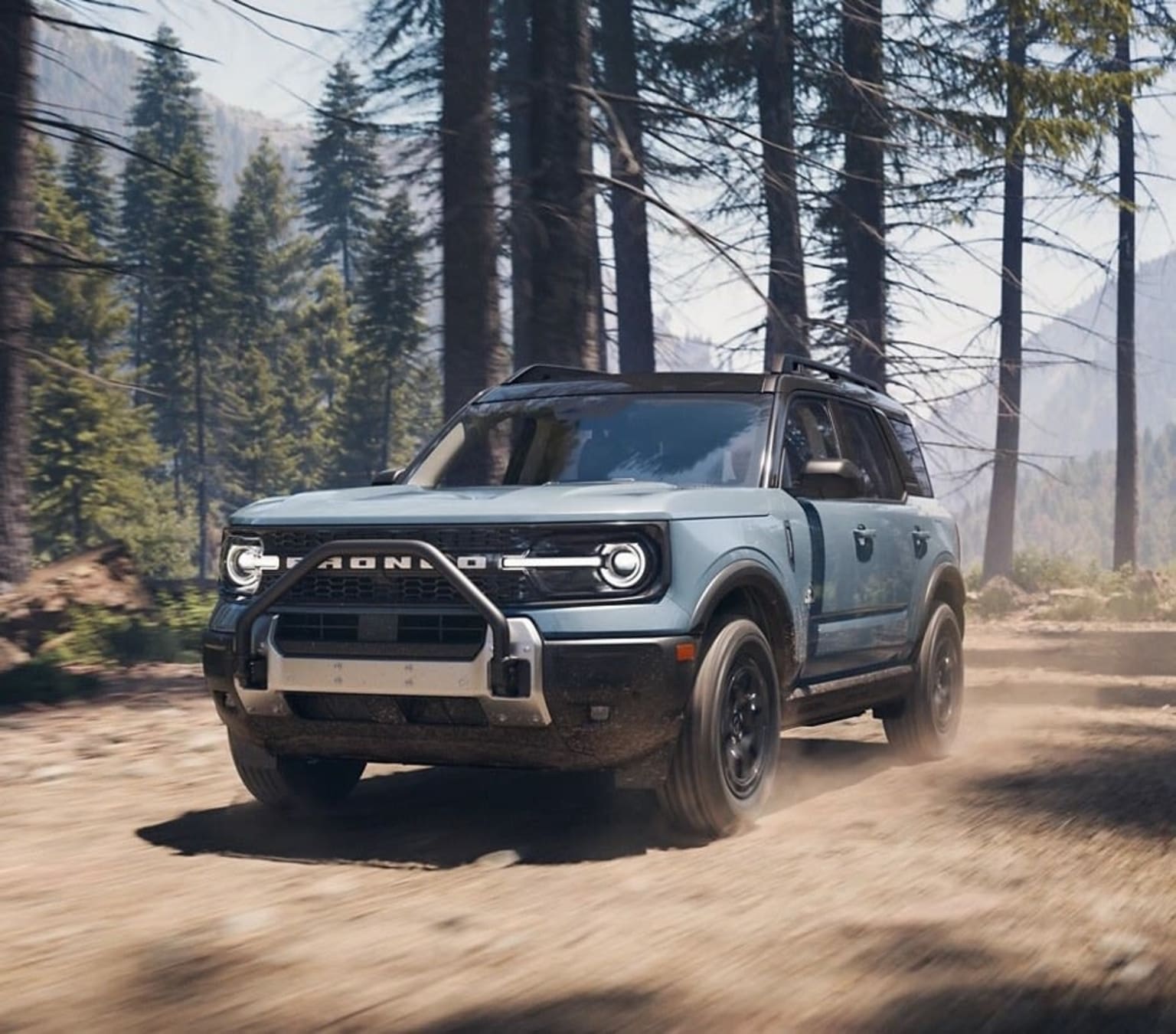 A light blue SUV drives on a dirt road in a forest with tall trees and mountains in the background.