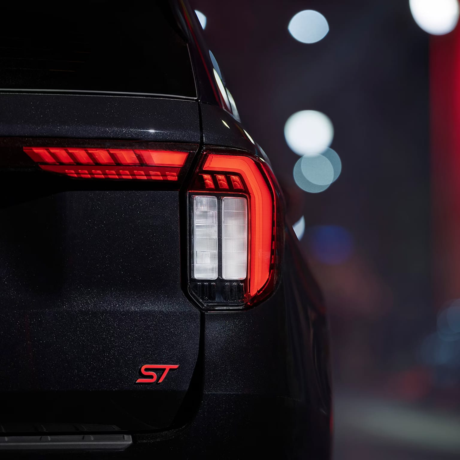 Close-up of a dark SUV's illuminated taillight with an "ST" badge in the foreground, set against a blurred background of bokeh lights.