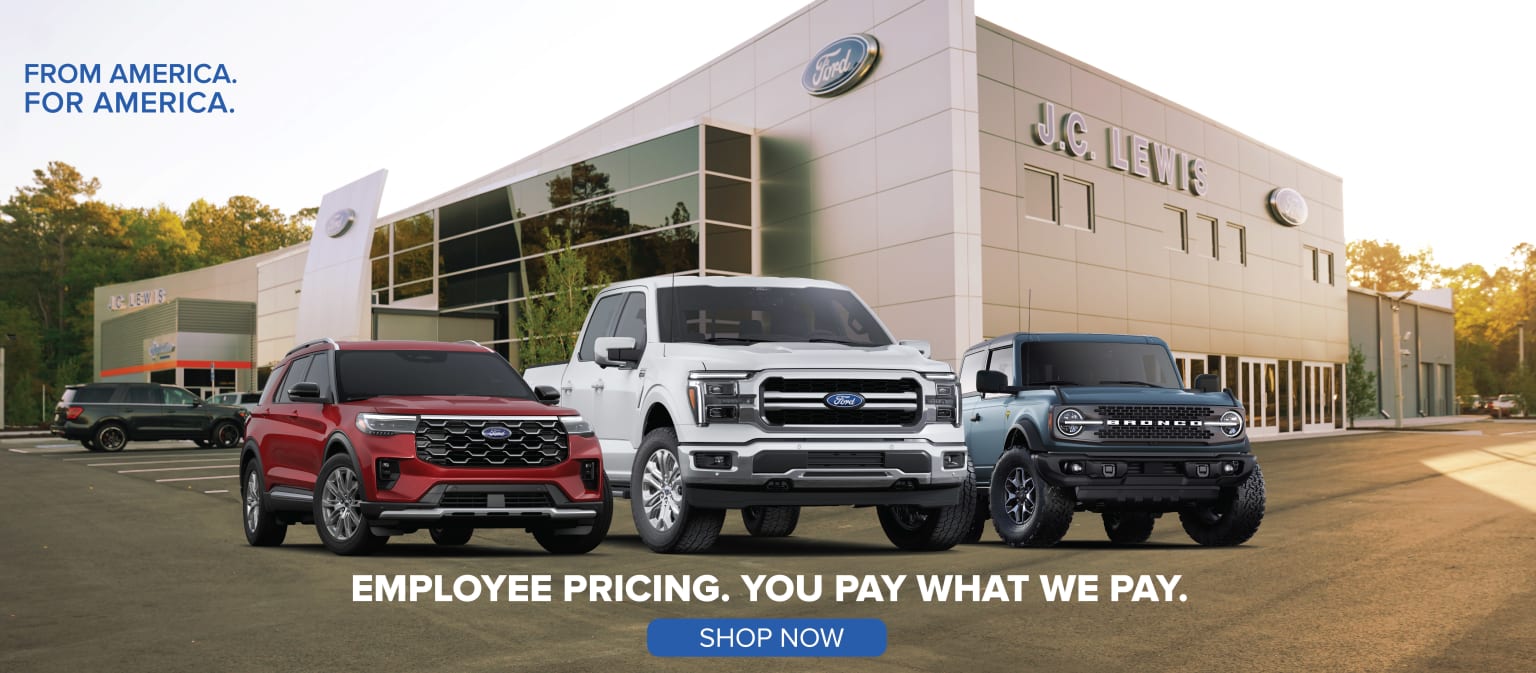 The image shows a car dealership with several large pickup trucks parked in the foreground, including a red and a white truck. The dealership building in the background has the company's logo prominently displayed.