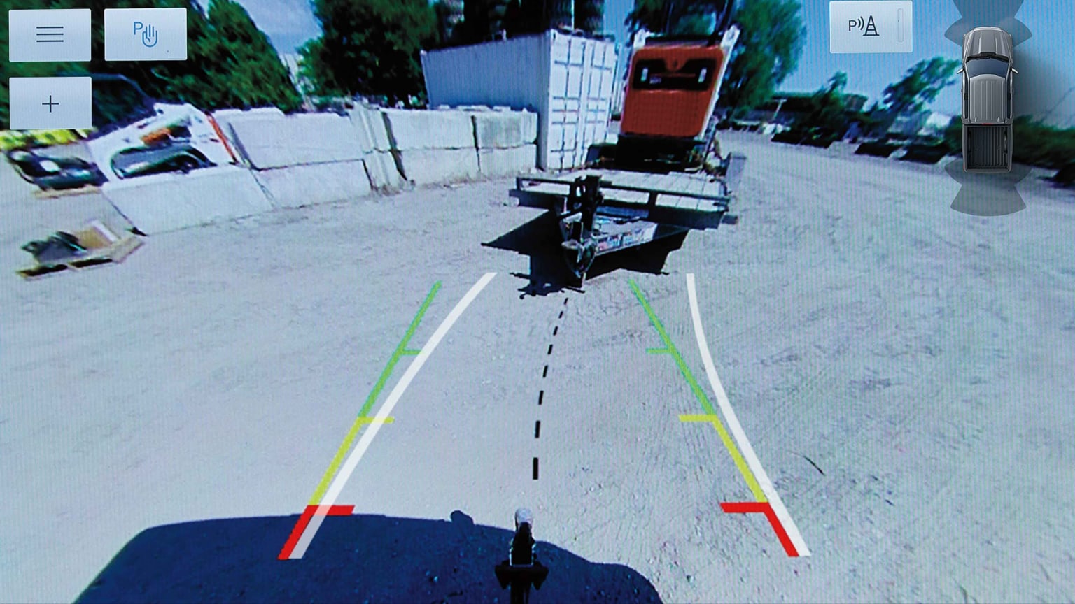 Backup camera view showing guiding lines on a sandy lot with a trailer and concrete blocks in the foreground and a truck and shipping container in the background.