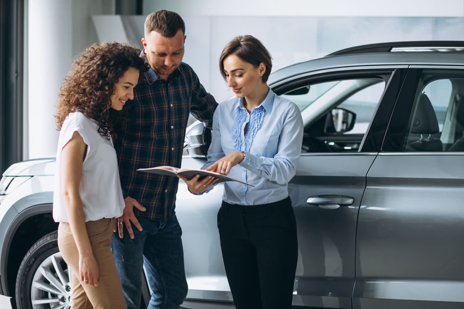 Image showing three people, two men, and one woman, discussing in a car dealership showroom. The woman is holding a tablet.