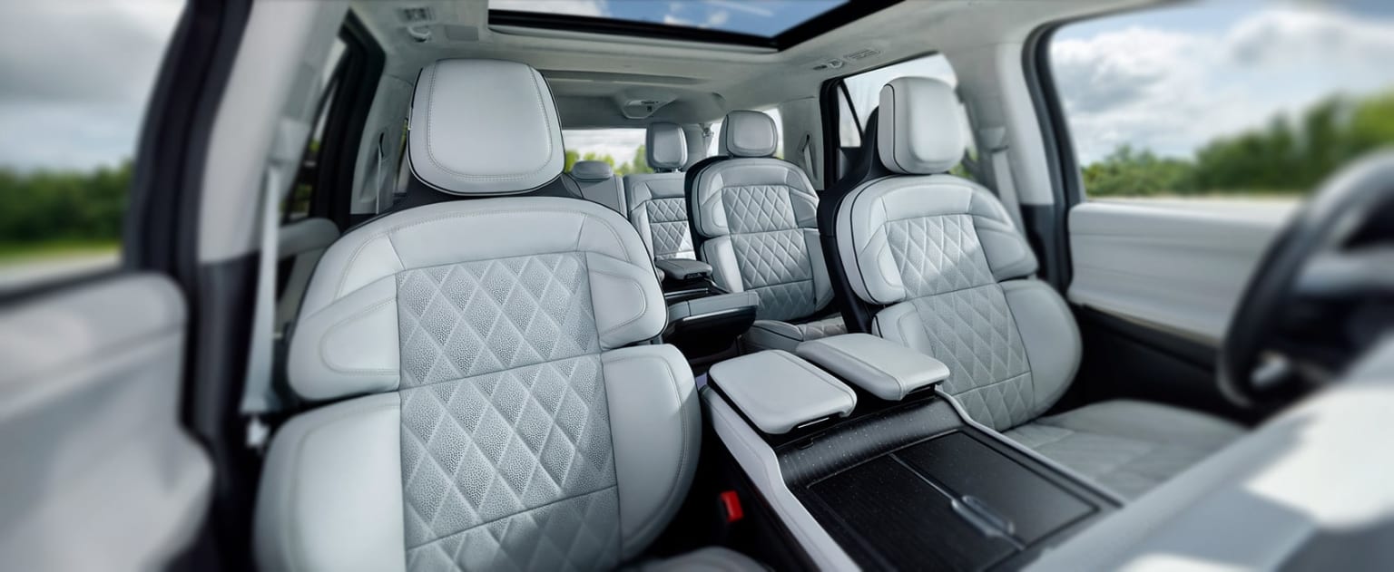 The image shows the interior of a vehicle, with a row of gray, quilted leather seats in the foreground and a view of the exterior through the windshield in the background.