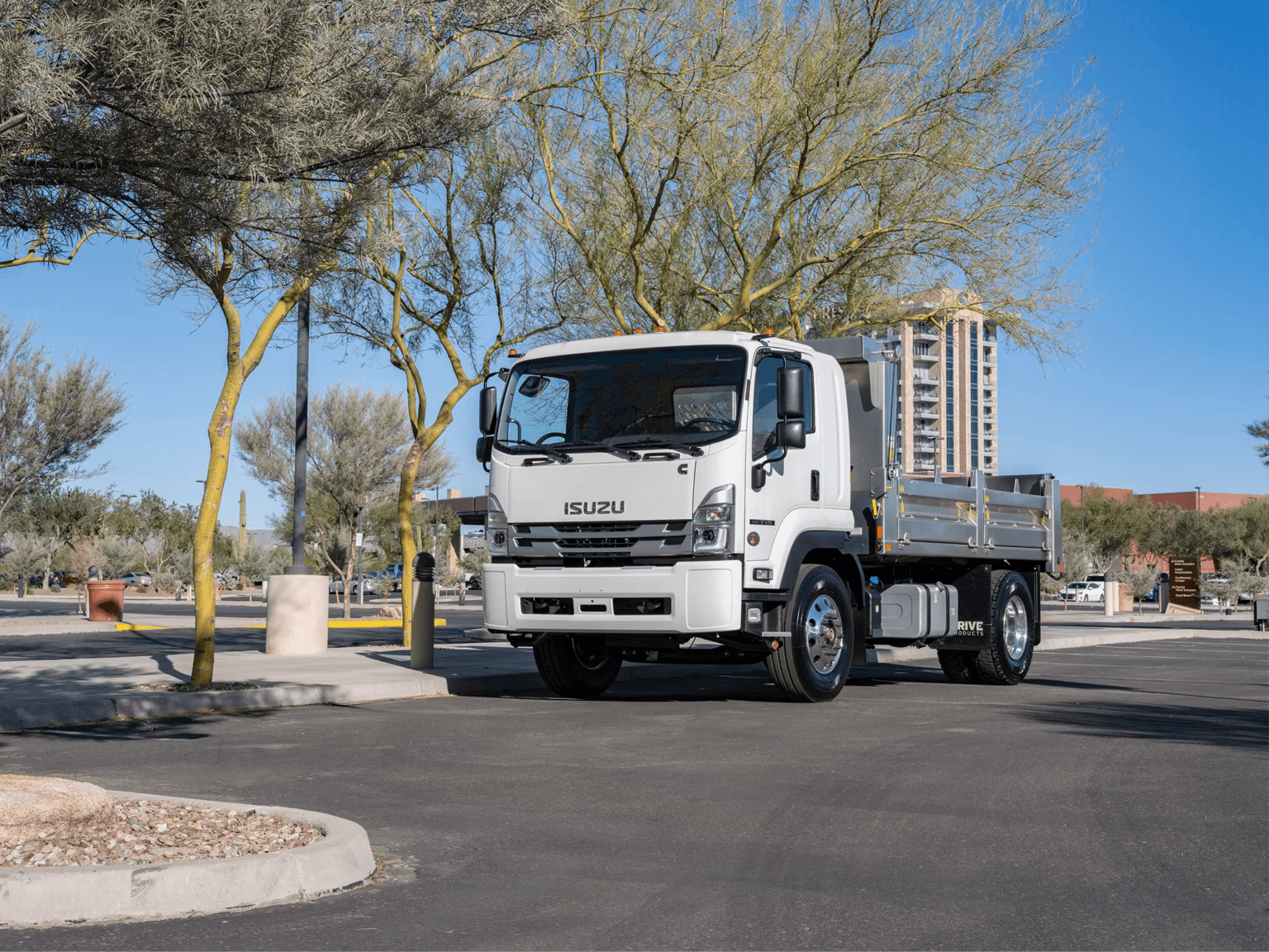 A white dump truck is parked on an asphalt lot, surrounded by sparse trees and a tall building in the background.