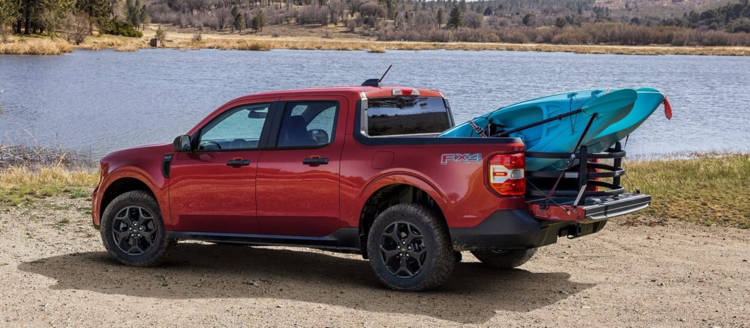 A red pickup truck with a kayak attached to the back parked on a sandy beach near a lake surrounded by forested hills.