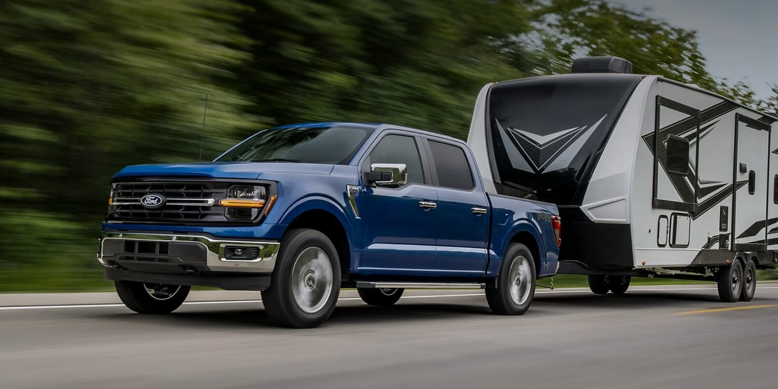 A blue pickup truck is towing a large recreational vehicle on a road surrounded by lush greenery.