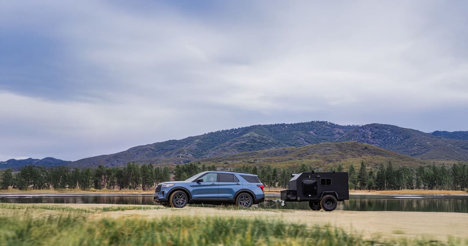 SUV parked next to a trailer in a grassy field with a mountainous landscape and cloudy sky in the background.