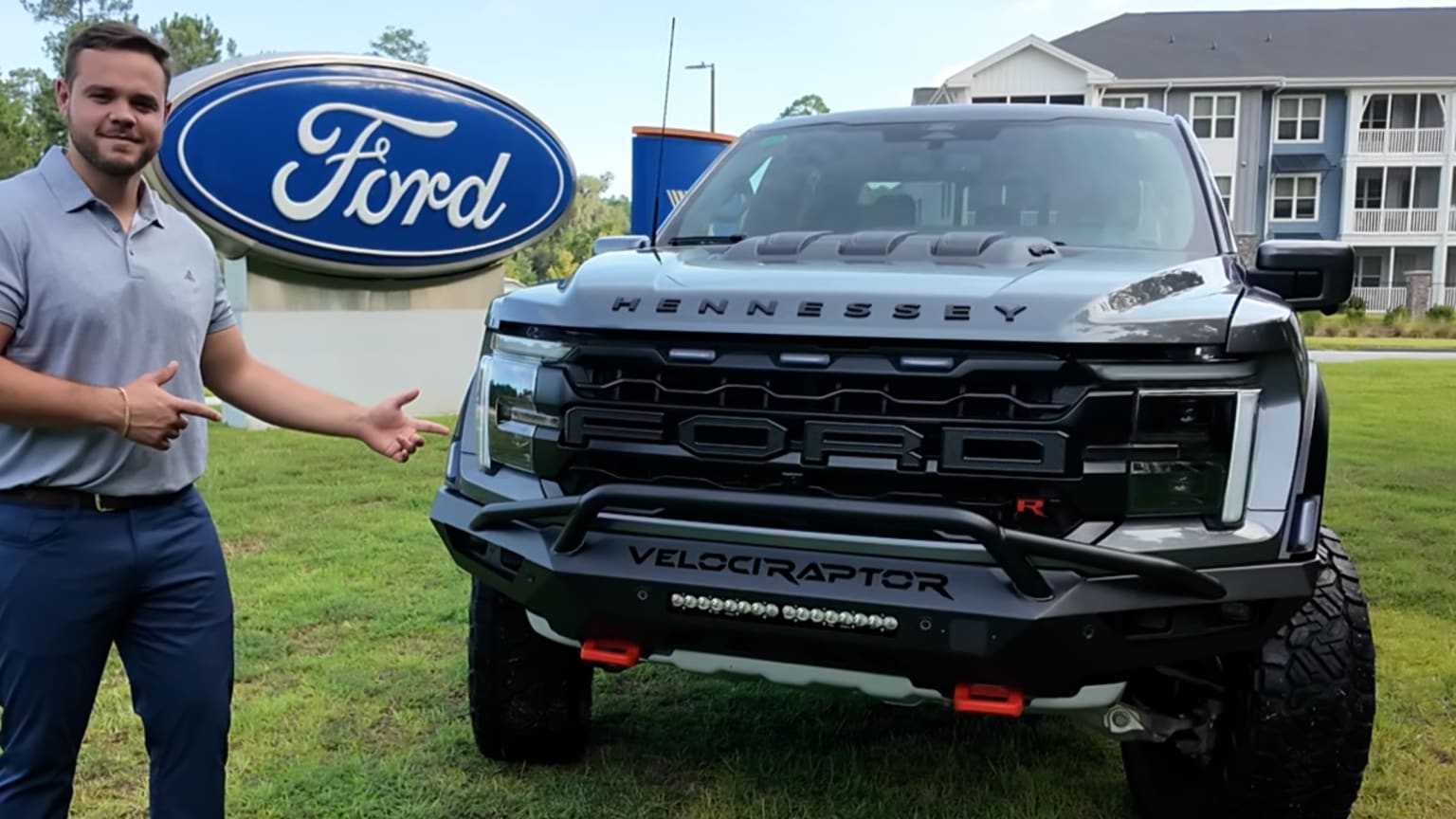 A man points to a modified truck parked in front of a dealership with a modern building in the background.