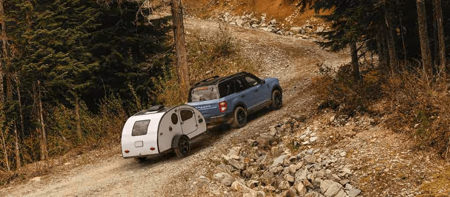 A blue SUV towing a small white teardrop trailer travels along a rocky dirt road surrounded by a dense evergreen forest.