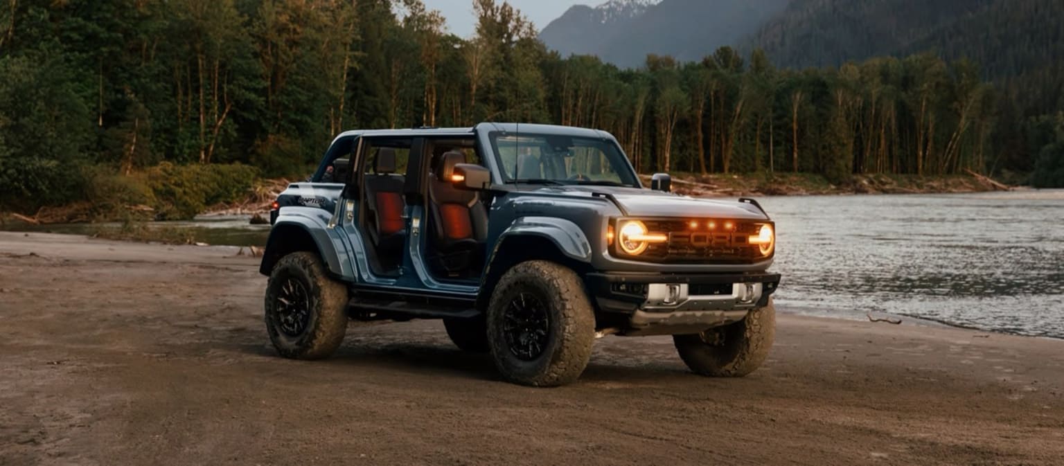 An off-road vehicle in a forested area beside a river with headlights on, set against a mountainous backdrop.