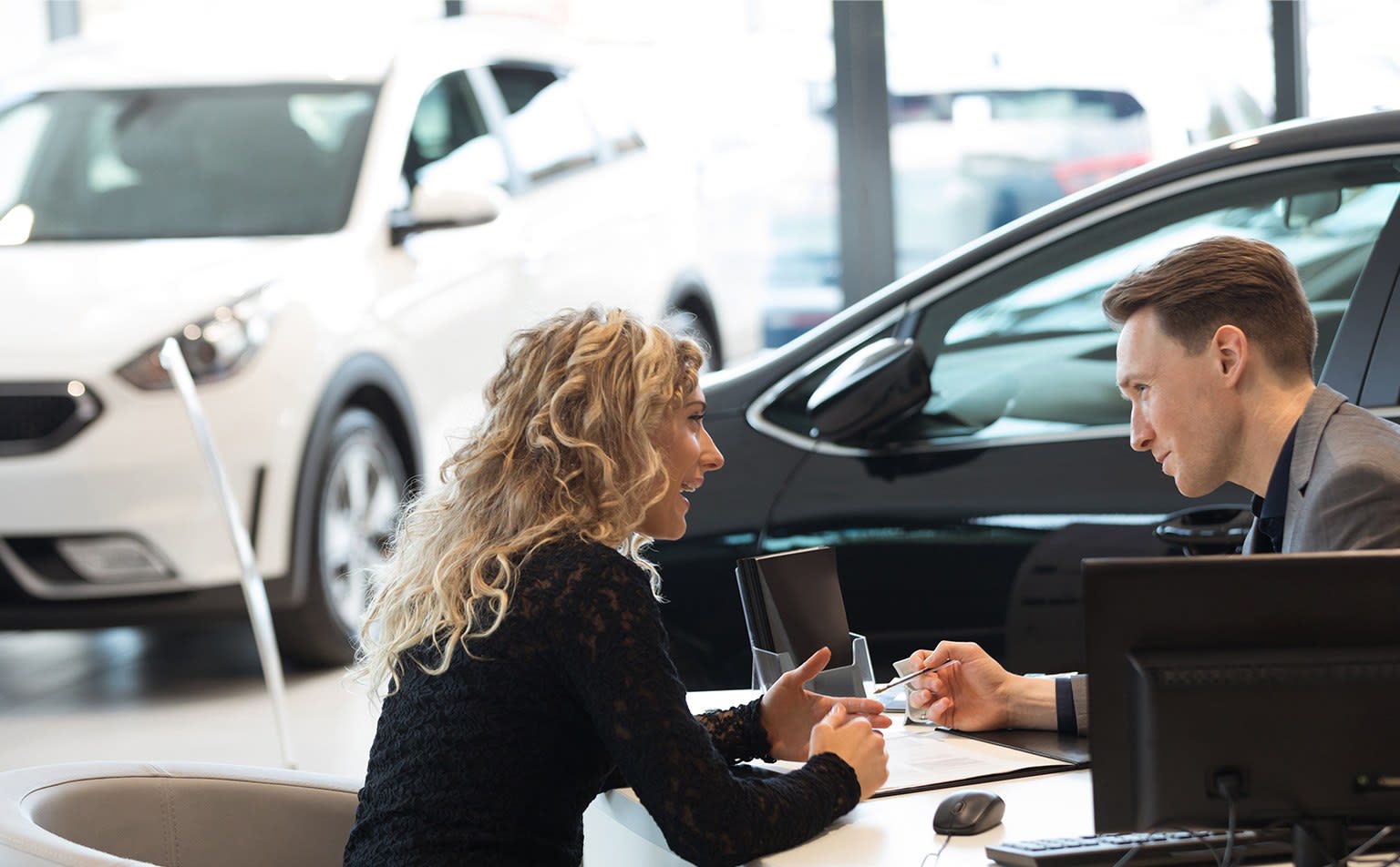 A woman with curly hair and a man talk in a car dealership showroom with cars in the background.