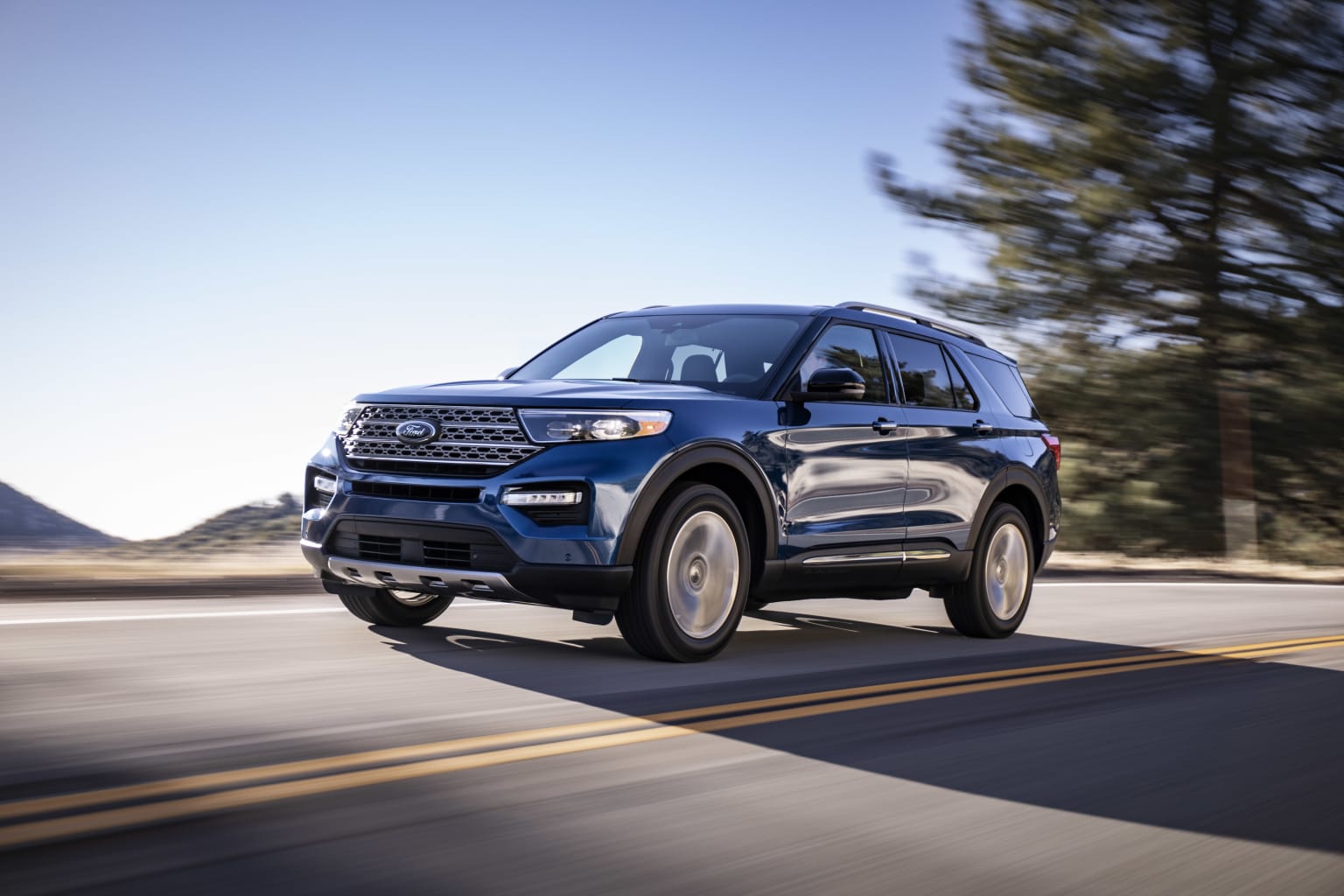 a blue ford explorer is driving down the road in front of a mountain and pine trees in the background