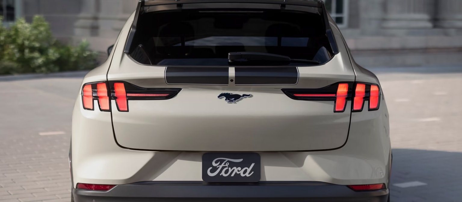 A white Ford Mustang SUV with distinctive taillights and a Ford logo on the rear, parked on a city street with buildings visible in the background.