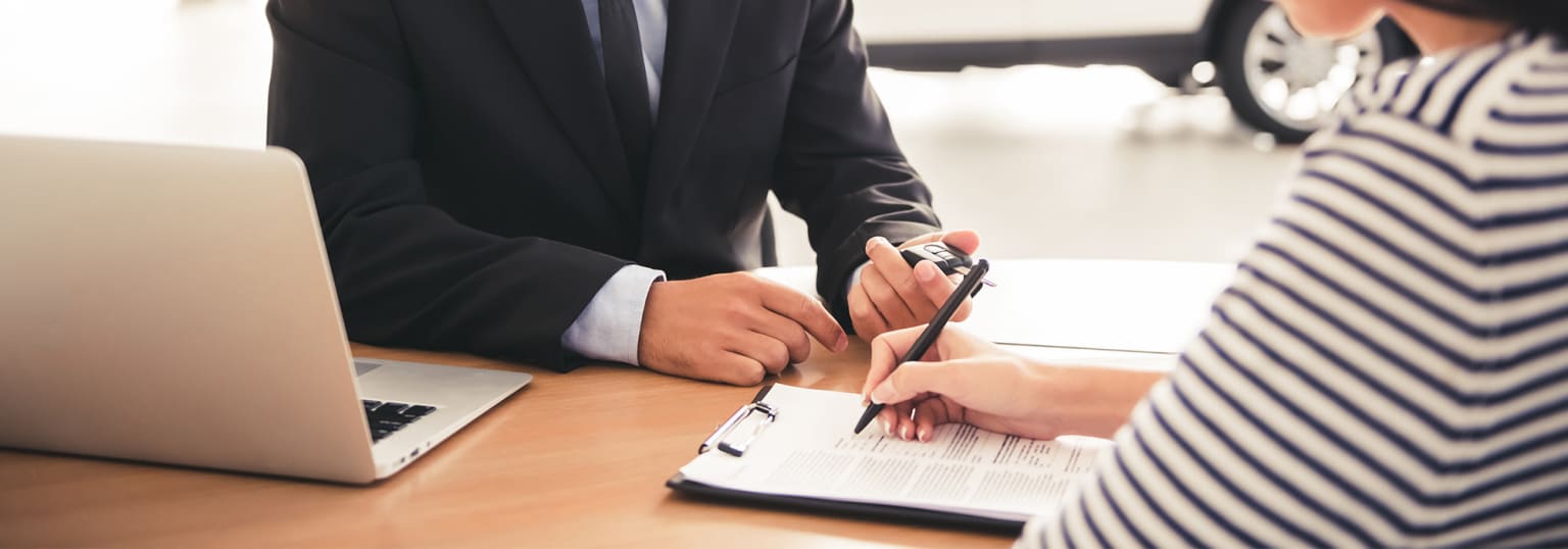 A person in a suit signing or reviewing documents on a desk with a laptop computer in the background.