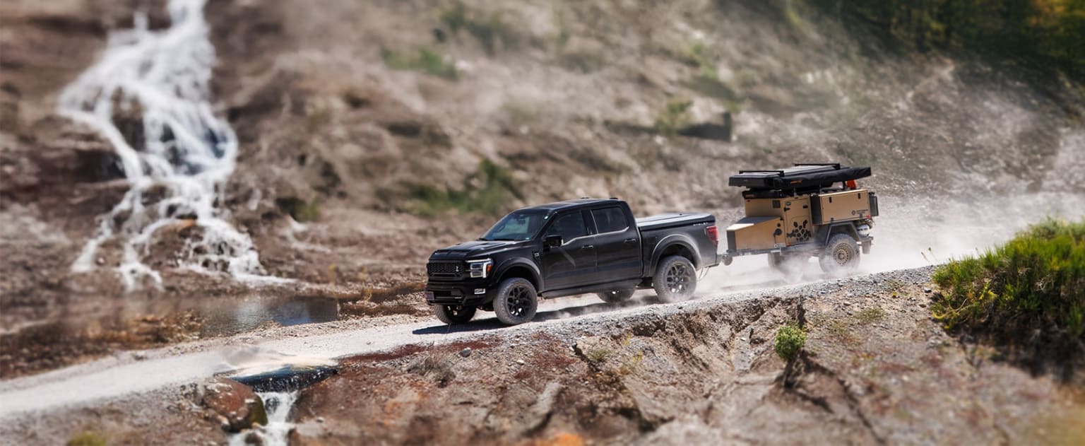 A black pickup truck with a trailer attached driving on a dirt road in a mountainous landscape with a waterfall in the background.