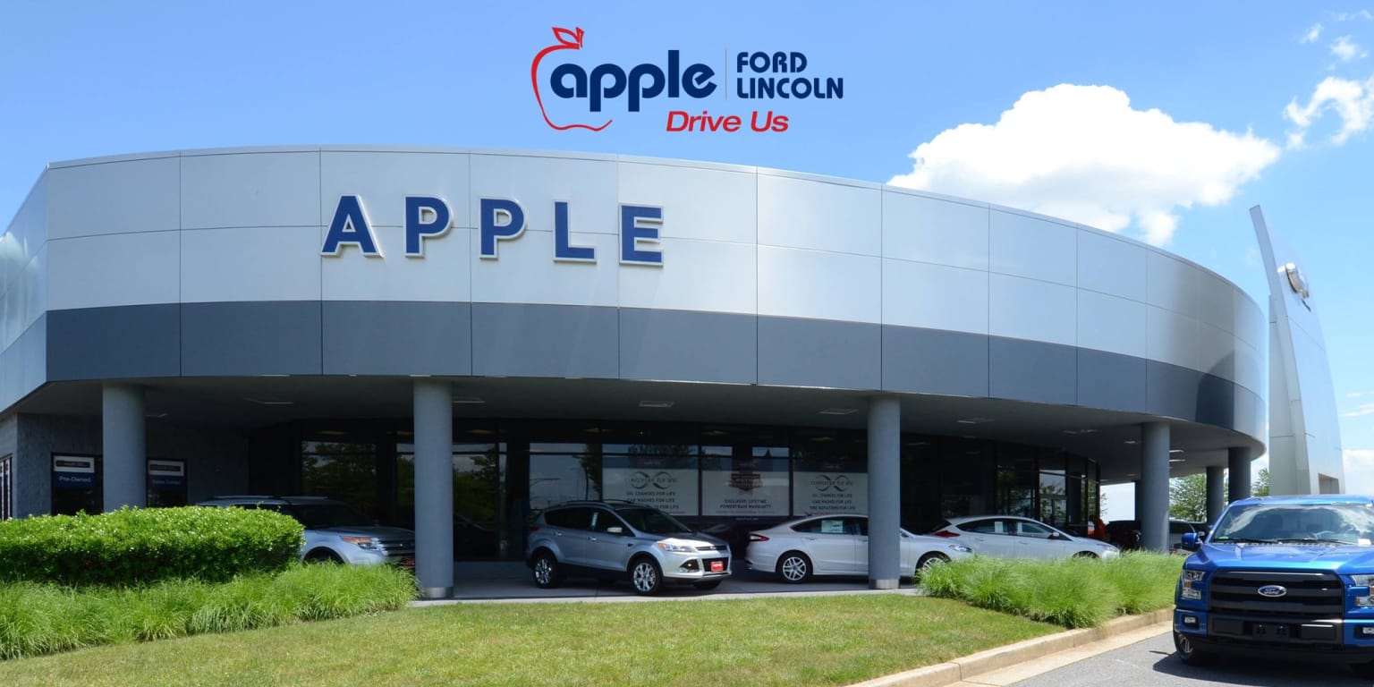 A modern car dealership with a curved facade and a blue pickup truck in the foreground sits under a bright blue sky with fluffy clouds.