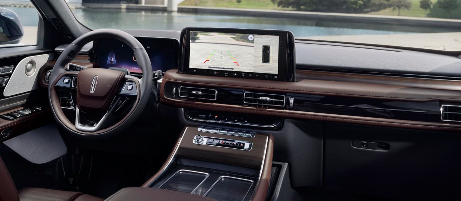 Luxurious car interior with a large touchscreen displaying a backup camera, with a calm body of water and a bridge in the background.