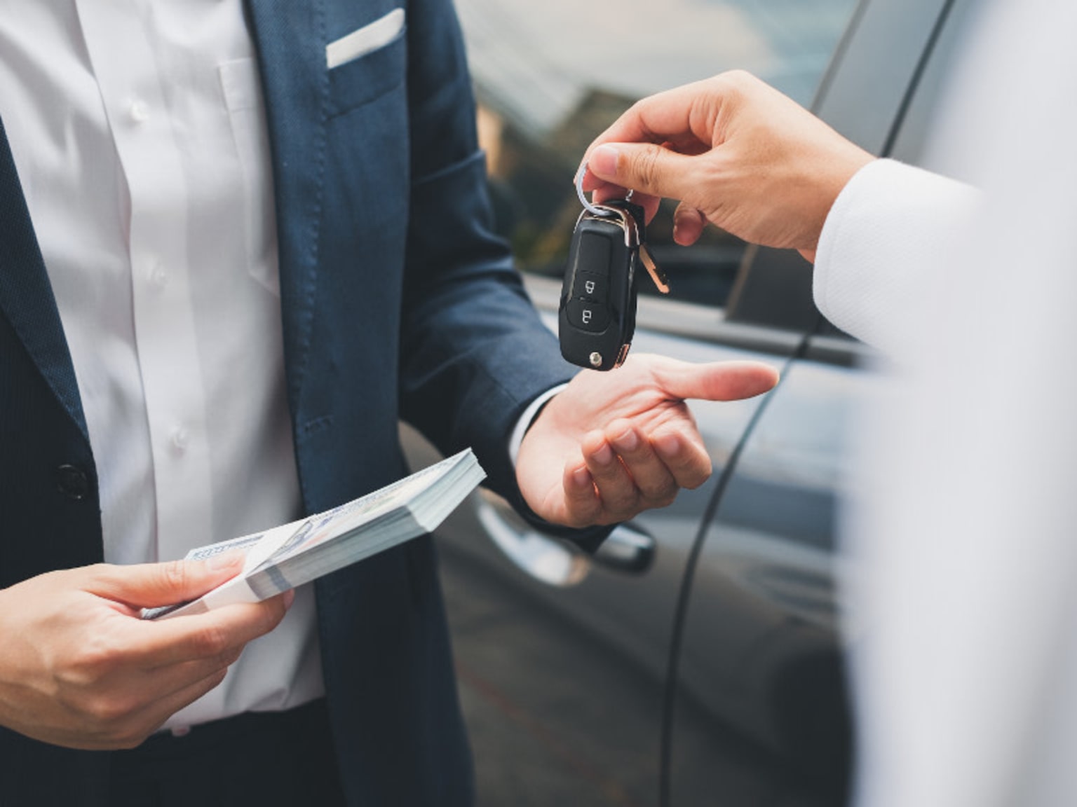 A person in a suit holding a car key and a document, standing next to a car.