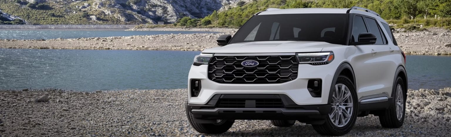 A white SUV parked on a rocky shore with a calm body of water and mountains in the background.