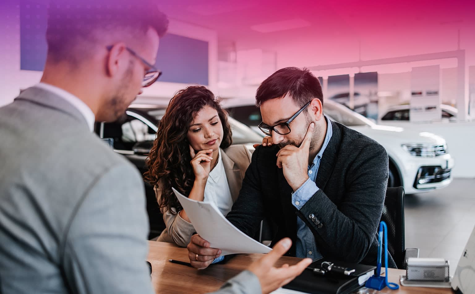 Three individuals examining a document in a car dealership showroom with various vehicles in the background.
