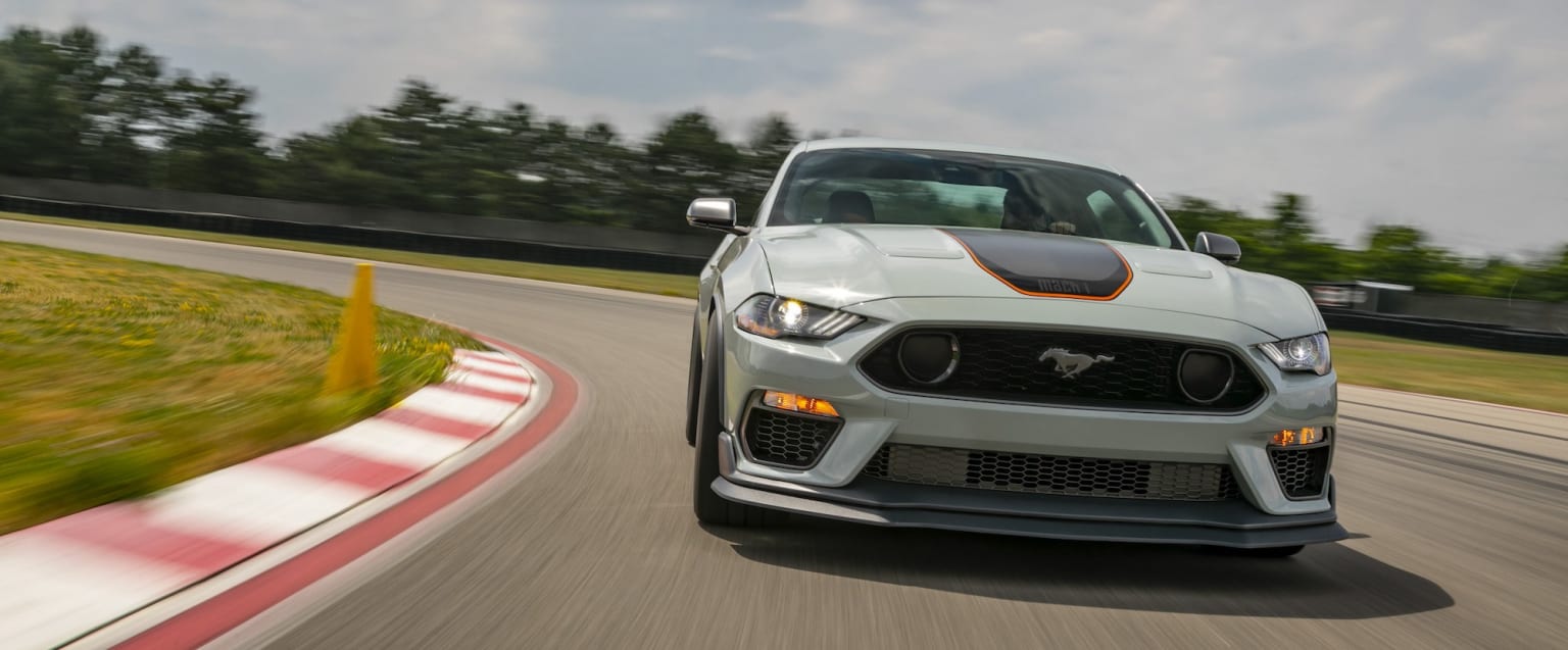 a white mustang mustang driving down a race track with trees in the background and a red and white stripe on the front of the car