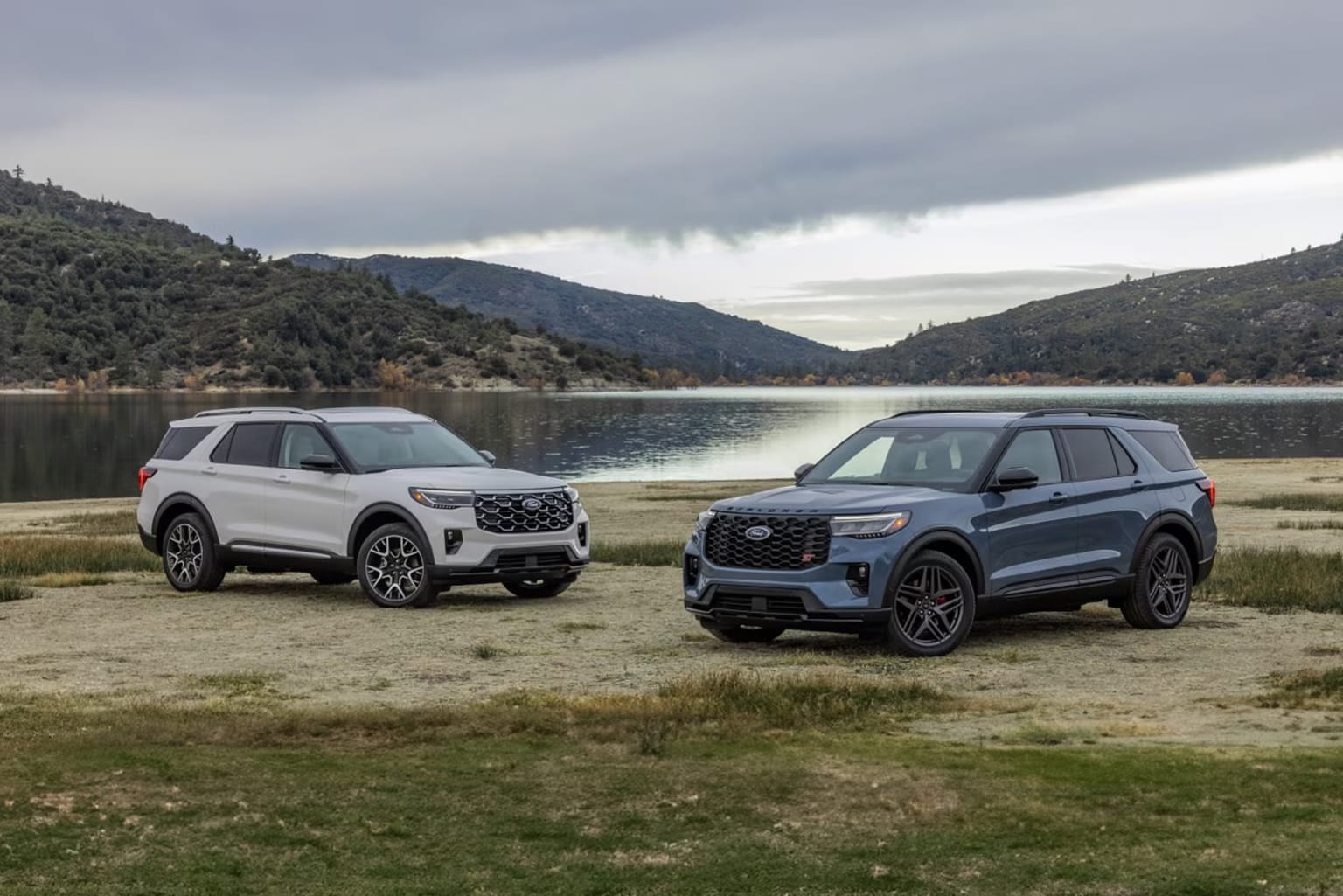 Two large SUVs parked on a grassy area near a serene lake surrounded by mountains and a cloudy sky.