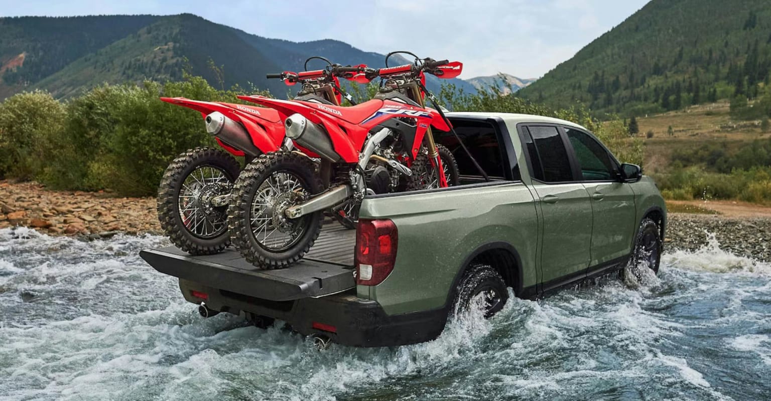 Two dirt bikes in the bed of a green pickup truck driving through a shallow river, surrounded by lush green mountains and trees.