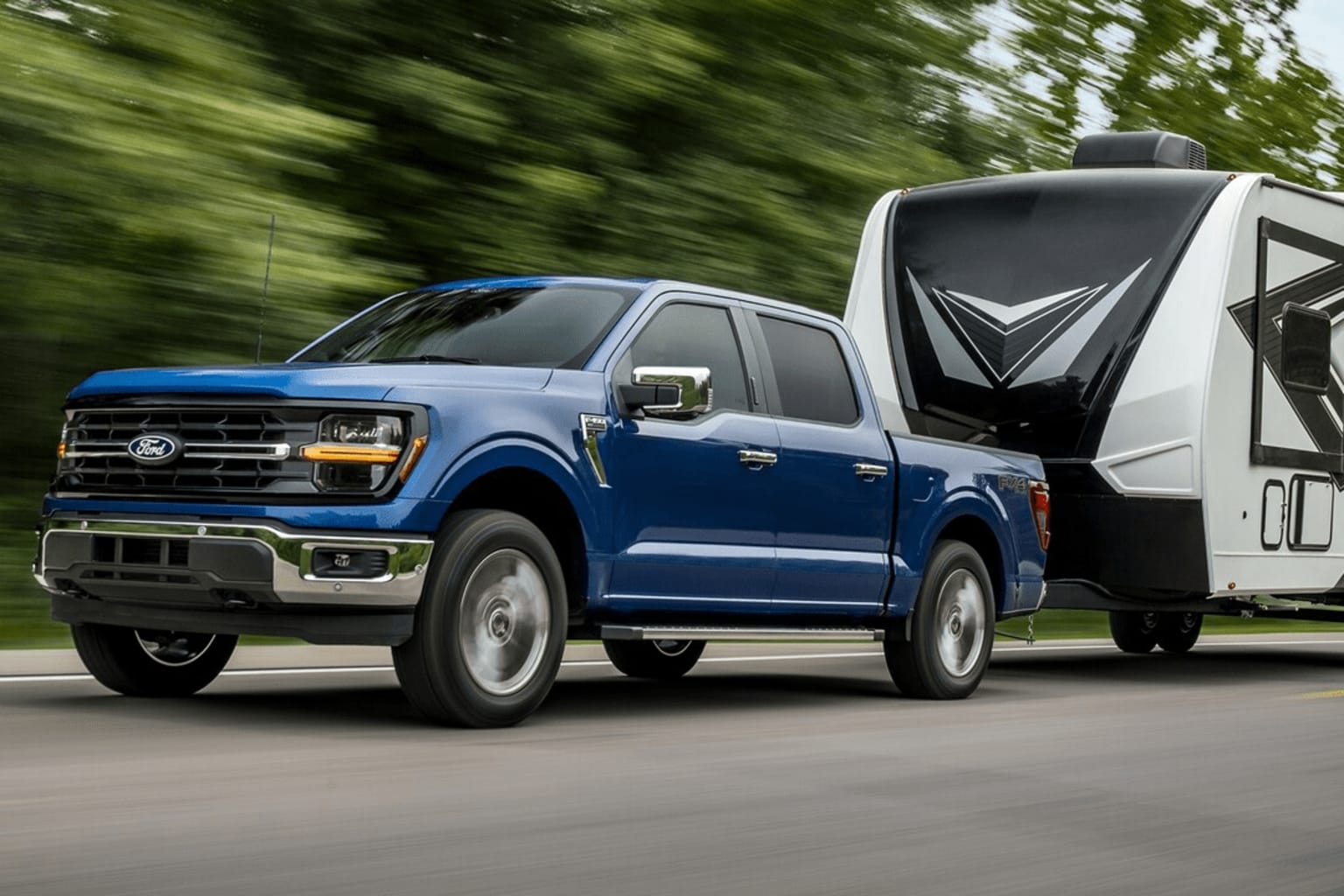 A blue pickup truck towing a large white recreational vehicle (RV) through a wooded area, conveying a sense of motion and speed.