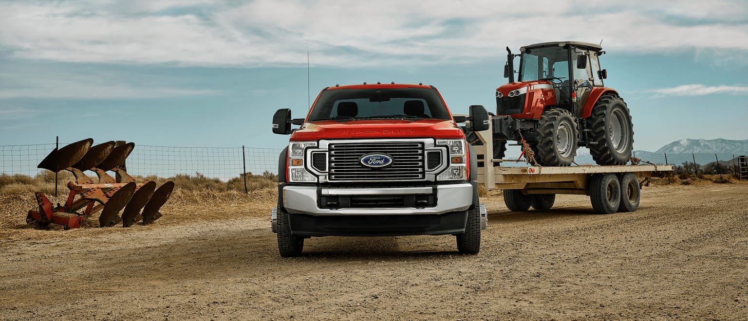 A large red and silver truck is transporting a red tractor on a dirt road, with a cloudy sky and a rural landscape in the background.