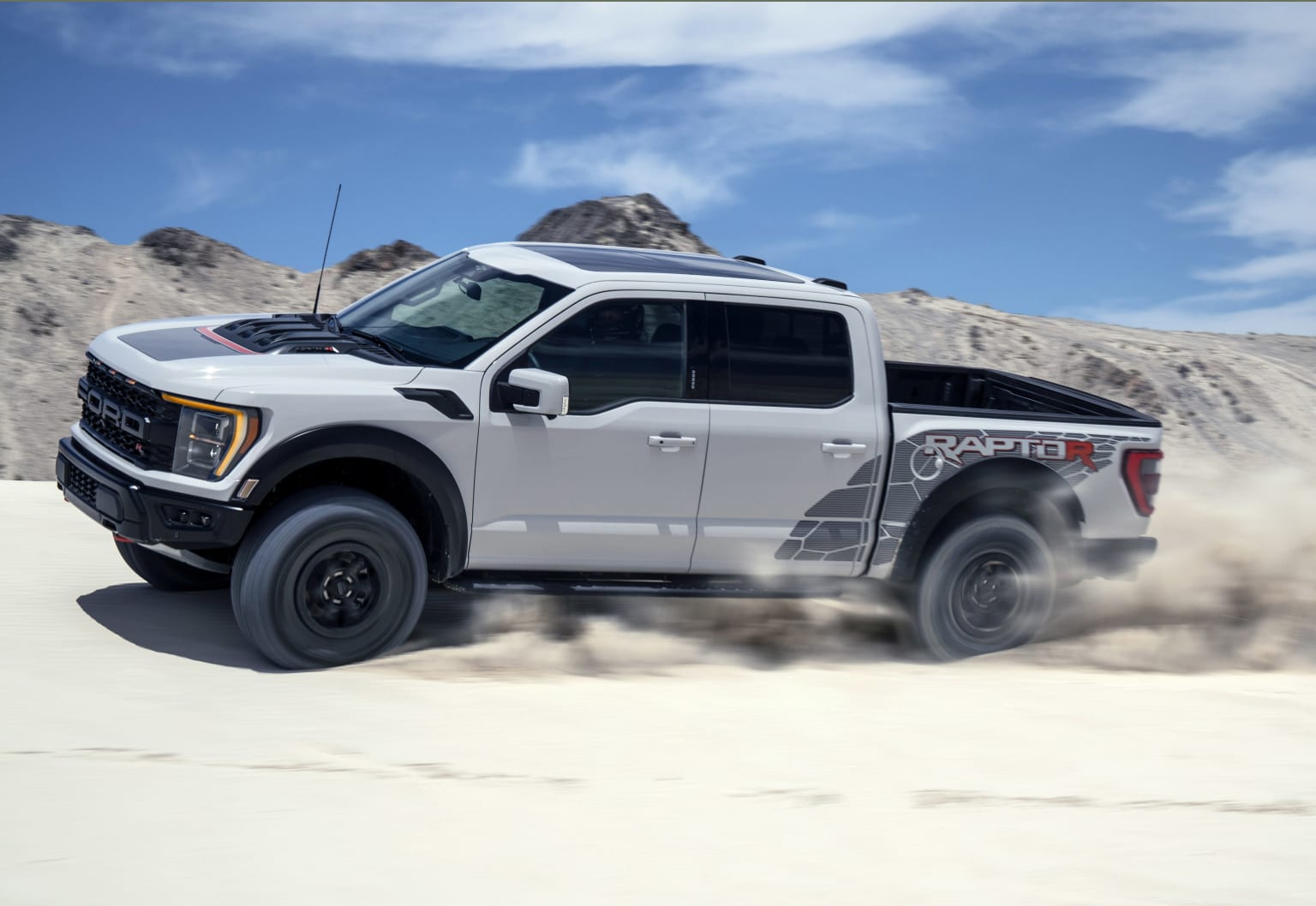 a white truck driving through a desert with mountains in the backgrouund and clouds in the sky