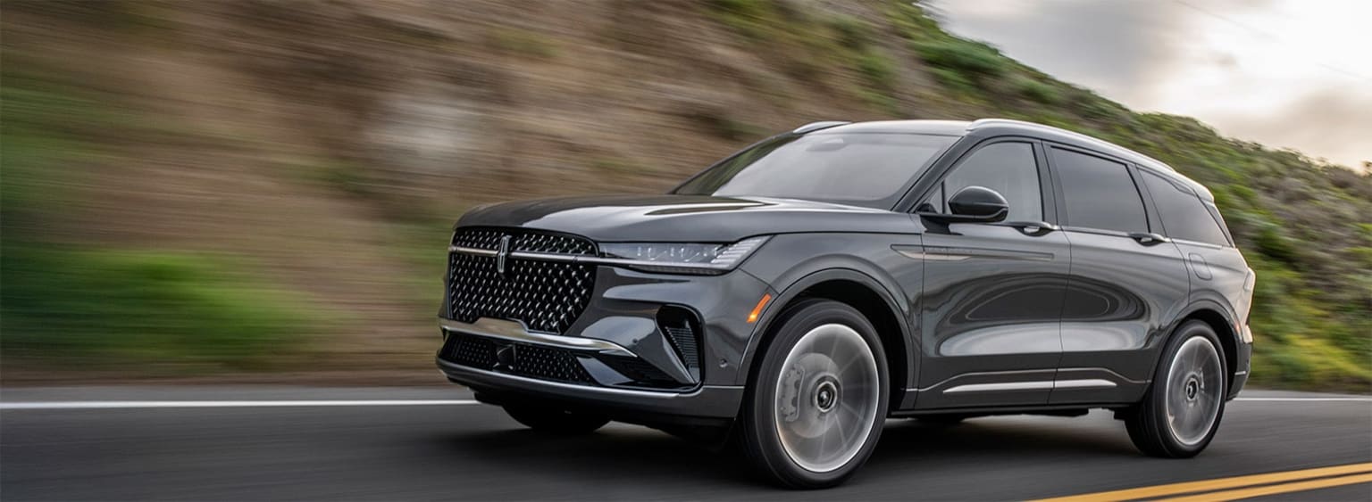 A sleek, gray SUV speeds through a blurred, forested landscape, its aggressive front grille and sharp lines conveying a sense of power and dynamism.