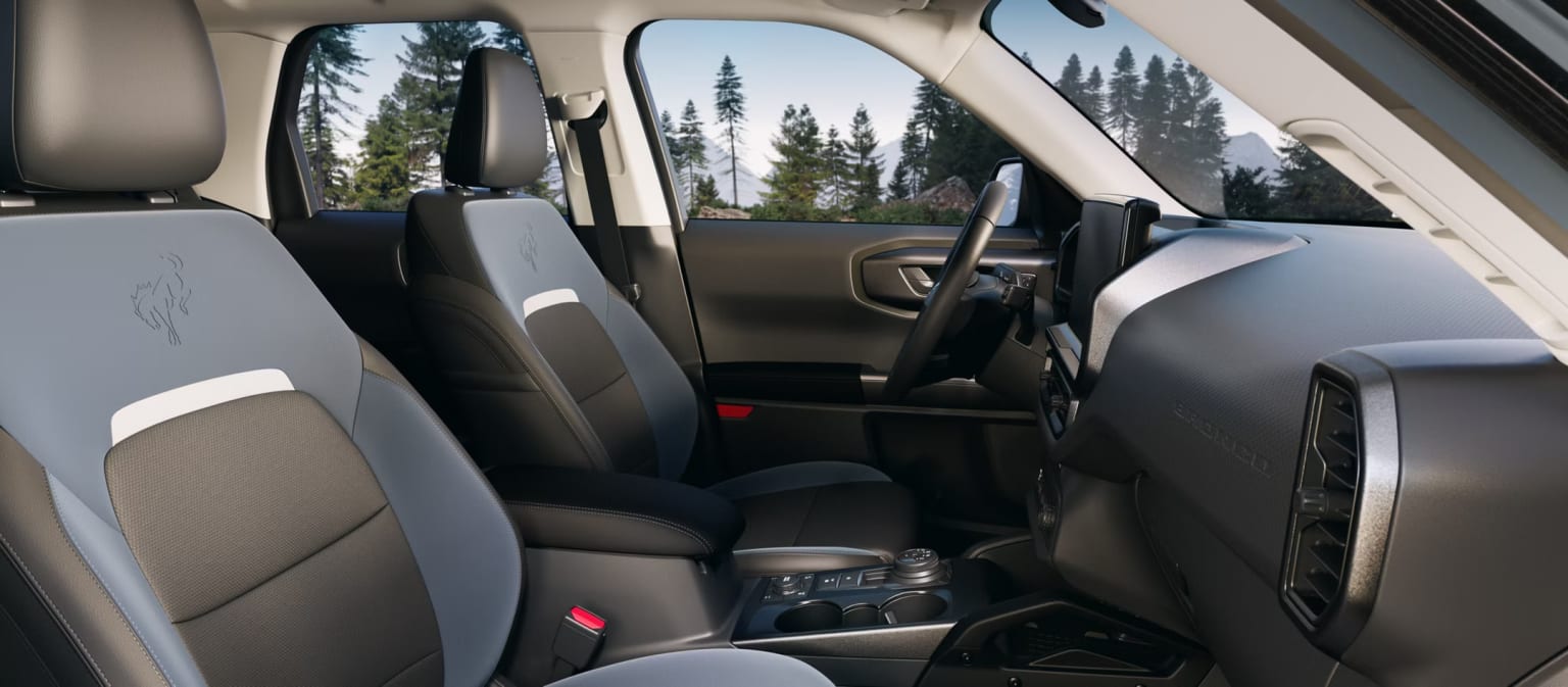 Interior view of a vehicle with front seats and dashboard, set against a background of a forested area with trees and a clear sky.
